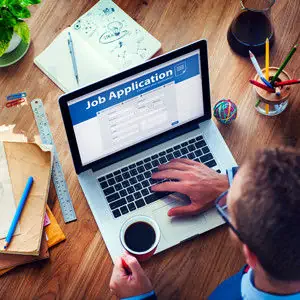 An image of someone sat at a desk, holding a cup of coffee and operating a laptop