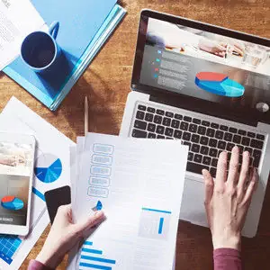 An image of someone holding papers over a desk, while operating a laptop